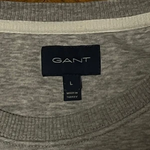 Grå Gant tröja - Snygg grå sweatshirt från GANT med rund hals och broderad logga samt sköld på bröstet. Tröjan har ribbade muddar vid ärmslut och nederkant, vilket ger en schysst passform. Perfekt för chill dagar och enkel att matcha med alla typer av jeans.