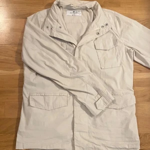 Ljusbeige field jacket John Henric - Snygg ljusbeige field jacket från John Henric med klassisk krage och flera fickor framtill. Jackan är tillverkad i bomull och har en stilren look. Använd fåtalet gånger men är lite skrynklig, kan lätt fixas. En perfekt jacka för hösten. Storlek S men sitter som M.  Nypris 1599, säljs nu för 650.