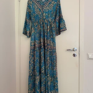 Blå långklänning med paisleymönster - Säljer en bohemisk långklänning med paisley- och blommönster i blått och beige. Klänningen har v-ringad hals, trekvartsärmar med lätt volang och är tillverkad i 80% konstsilke och 20% polyester. Perfekt för dig som gillar färgstarka och unika plagg.