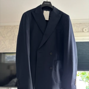 Mörkblå kavaj från These Glory Days - Snygg mörkblå ny kavaj från These Glory Days med klassisk passform och dubbla knappar framtill. Kavajen har breda slag, innerficka och slät yta. Fodret är i mörkblått satinliknande material och knapparna är svarta. Perfekt för dig som gillar stilrena och tidlösa plagg. Strl M