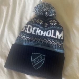 Blå DIF mössa  - Snygg blå mössa med stor tofs på toppen och broderad DIF-logga framtill. Mössan har mönster med snöflingor och zigzag i vitt och ljusblått samt texten 'Stockholm' runt om. Perfekt för dig som vill visa ditt stöd för Djurgården under kalla dagar.