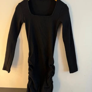 Svart klänning  - Svart klänning med långa ärmar och fyrkantig halsringning. Klänningen har rynkdetaljer på sidorna och sitter tajt. Storlek S
