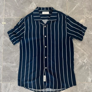 Blåvit randig skjorta från Pier One - Snygg kortärmad skjorta från Pier One i marinblått med vita vertikala ränder. Skjortan har klassisk krage, knappar framtill och en avslappnad passform. Perfekt för dig som gillar stilrena och fräscha plagg med lite edge.