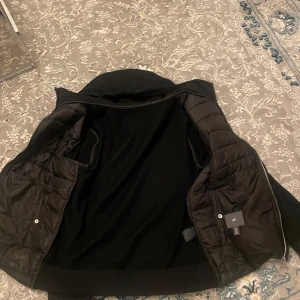 Svart pufferjacka med huva från H&M - Svart pufferjacka från H&M med quiltad framsida och mjuka ärmar. Jackan har huva med snörning, två fickor med tryckknappar och dragkedja framtill. Perfekt för dig som gillar en clean och chill stil. Passar dig som vill ha en jacka med lite extra edge.