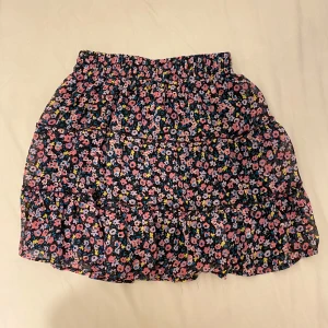 Blommig volangkjol från FB Sister (new Yorker) - Supersöt kort kjol från FB Sister med blommigt mönster i rosa, lila, blått och gult på svart botten. Kjolen har volanger och resår i midjan för en skön passform. Perfekt för dig som gillar färgglada och lekfulla plagg. Använd typ 2 gånger 