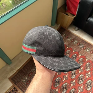 Svart Gucci keps med GG-mönster - Snygg svart keps från Gucci med klassiskt GG-mönster över hela kepsen. Har en grön och röd rand på sidan och justerbar rem bak i skinn. Perfekt för dig som vill ha en lyxig och stilren accessoar med street vibe.
