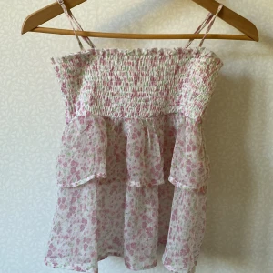 Blommig volangtop från Gina Tricot - Söt blommig topp från Gina Tricot med volanger och smock upptill. Toppen har tunna axelband och är tillverkad i ett lätt, luftigt material med rosa och gröna blommor på vit botten. Perfekt för sommarens alla häng och picknickar.