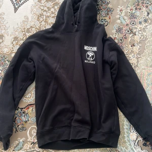 Svart Moschino Milano hoodie - Svart hoodie från Moschino Milano med stor vit logotyp på ryggen och mindre tryck på bröstet. Tröjan har huva med snörning och långa ärmar. Mjuk och skön bomullskänsla, perfekt för chill eller streetwear. Snyggt och stilrent statement-plagg.