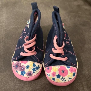 Blommiga baby sneakers med kattmotiv - Supersöta baby sneakers i marinblå bomull med rosa snörning och blommigt mönster på tå och sula. Sidorna har broderat kattansikte med rosett. Platt sula med små tassavtryck undertill. Perfekta för små äventyrare som gillar färg och gulliga detaljer. Står ej för frakten 