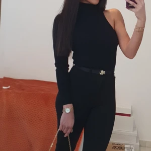 Svart oneshoulder polotopp - Snygg svart polotopp med oneshoulder-design och lång ärm på ena sidan. Toppens tighta passform ger en clean och trendig look, perfekt för dig som gillar minimalistisk stil. Materialet är mjukt och stretchigt, vilket gör den bekväm att bära hela dagen.