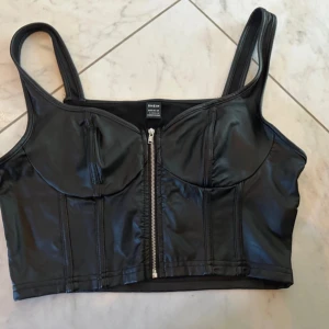 Svart korsett-topp med dragkedja SHEIN - Cool svart korsett-topp från SHEIN med breda axelband och snygg dragkedja framtill. Topen har en croppad passform och är gjord i glansigt syntetmaterial som ger en edgy vibe. Perfekt för att lyfta din outfit med lite attitude.
