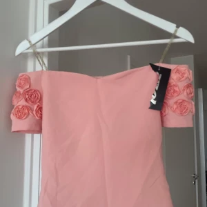 Rosa topp med rosdetaljer - Supersöt ljusrosa kort topp från i storlek 36. Klänningen har offshoulder-ärmar dekorerade med tygrosor .