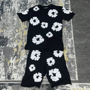 Svart blommig t-shirt från Denim Tears - Svart t-shirt från Denim Tears med stora vita blommönster över hela plagget. T-shirten har rund hals och korta ärmar, tillverkad i mjuk bomull. Perfekt för dig som gillar statement-plagg och vill sticka ut med cool design.