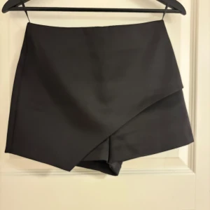 Svarta omlottshorts från Topshop - Snygga svarta shorts från Topshop i omlottmodell med stilren design och dragkedja bak. De är högmidjade och har en clean look som passar perfekt till en trendig outfit. Materialet känns mjukt och har en lätt glansig finish.