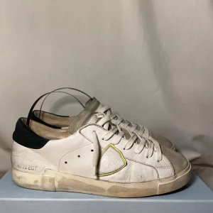 Philippe Model vita sneakers - Snygga vita sneakers fràn Philippe Model med grà mockadetaljer pả tän och svart häl. Skorna har snörning, rund tà och en ikonisk sköldlogga pả sidan med gulgrön kant. Ovandelen är i skinn och mocka, perfekt för en avslappnad streetstil.