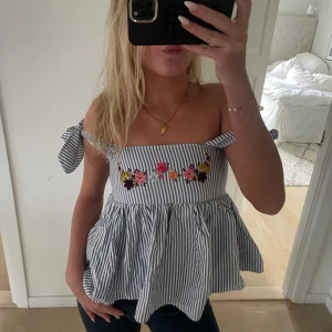 Randig blommig blus med knytband - Supersöt blus med blåvita ränder och broderade blommor framtill. Modellen har volang nedtill och knytbara axelband som kan bäras offshoulder eller med axelband. Luftig och flowy passform, perfekt för varma dagar.