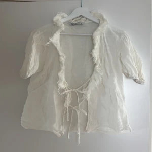 Vit linne blus med volang och knyt från Zara - Säljer en söt vit blus från Zara med korta puffärmar och volangkant längs den öppna fronten. Blusen har två knytband framtill och är gjord i ett lätt, luftigt linnematerial.  vibe.
