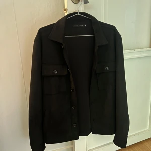 Svart mocka overshirt från Brand Island - Svart overshirt från Brand Island i mjuk mocka med klassisk krage och bröstfickor med knappar. Jackan har lång ärm och knäppning framtill, perfekt för lager-på-lager och en clean look. 