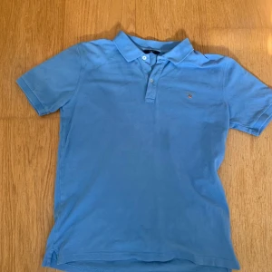 Blå pikétröja från Gant - Snygg blå pikétröja från Gant med klassisk krage och två knappar framtill. Tröjan har korta ärmar och en liten broderad logga på bröstet. Perfekt för dig som gillar stilrena och sportiga plagg. Tillverkad i mjuk bomull som är skön mot huden.