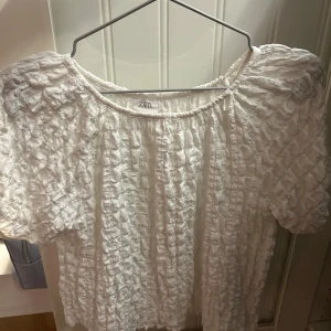 Vit blus med puffärm från Zara - Säljer en vit blus från Zara med korta puffärmar och ett coolt, krusigt tyg som ger en unik struktur. Blusen har rund hals och är perfekt för dig som gillar trendiga detaljer och en luftig känsla. Passar till jeans eller kjol för en fräsch look.