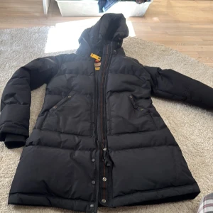 Svart dunjacka Masterpiece core parajumper XS - Svart dunjacka från Masterpiece Core i storlek XS. Jackan har huva med spänne, två stora fickor med knappar och cool patch på ärmen. Puffad design och dragkedja framtill. Perfekt för kalla dagar och har en riktigt schysst streetstil. Ny modell 