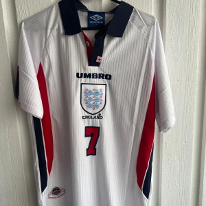 England Beckham 7 fotbollströja Umbro - Säljer en klassisk England fotbollströja från Umbro med nummer 7 och Beckham på ryggen. Tröjan är vit med tunna vertikala ränder, röda och marinblå detaljer på sidorna och kragen. England-emblem på bröstet och tryckta loggor. Perfekt för fans av landslaget.