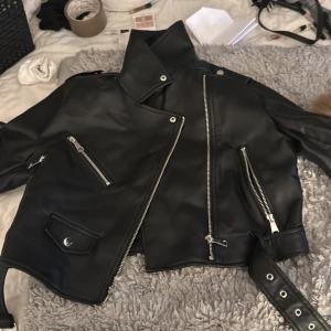 Svart skinnjacka från Stradivarius - Säljer en svart skinnjacka från Stradivarius med coola silverfärgade dragkedjor, snedställd front och bälte i midjan. Jackan har flera fickor med dragkedja och en klassisk bikerstil som ger en edgy vibe. Perfekt för dig som vill ha en snygg och trendig look.