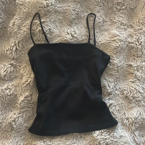 Svart ribbad linnetopp från KappAhl - Svart ribbad linnetopp från KappAhl med smala axelband och rak halsringning. Toppen är figurnära och har en enkel, stilren look som funkar till många olika outfits. Materialet är mjukt och stretchigt, perfekt för dig som gillar minimalistisk stil.