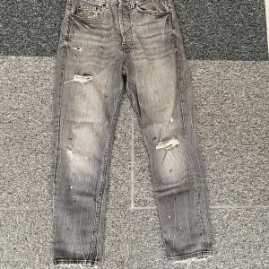 Grå jeans med slitningar från Pull&Bear - Säljer ett par grå jeans från Pull&Bear med coola slitningar och färgstänk i blått och vitt. Modellen har klassisk femficksdesign, raka ben och råa kanter vid benslut. Perfekt för dig som gillar en edgy streetstil och vill sticka ut lite extra.