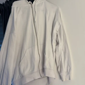 Vit hoodie med dragkedja från BikBok - Säljer en vit hoodie från BikBok i storlek XS. Tröjan har huva med snörning, dragkedja framtill och två stora fickor. Tillverkad i mjuk bomull, perfekt för chill dagar. Passformen är loose och ärmarna är långärmade. Enkel och clean stil.