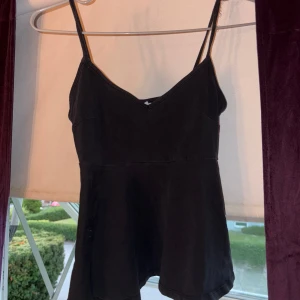 Svart linnetopp med v-ringning - Svart linnetopp med smala axelband och snygg v-ringning. Toppen har en lätt utsvängd nederdel som ger en cool siluett. Materialet känns mjukt och är troligtvis bomull eller polyester. Perfekt att styla med jeans eller kjol för en chill look. Urvättad och använd.