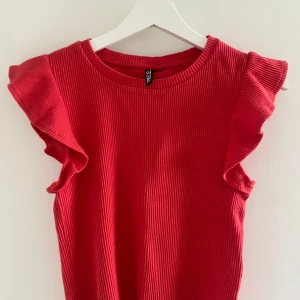 Röd ribbad topp med volangärmar - Snygg röd ribbad topp från PIECIES med volangdetaljer på axlarna. Modellen är ärmlös och har en rund halsringning. Perfekt för dig som vill ha en färgstark och trendig look med lite extra edge.