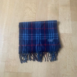 Blårutig kashmirhalsduk från Burberry - Snygg halsduk från Burberry i 100% kashmir, tillverkad i England. Mörkblå botten med rutigt mönster i ljusblått och röda linjer. Klassisk design med fransar i ändarna och mjuk känsla. Perfekt för att lyfta din stil under kyliga dagar.