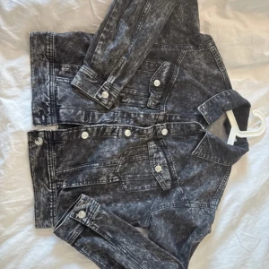 Svart tvättad jeansjacka med knappar - Snygg svart jeansjacka med tvättad look och vita knappar. Klassisk modell med krage, två bröstfickor med lock och långa ärmar. Jackan har en cool, lite sliten finish som ger den en edgy vibe och passar perfekt till streetwear-stilen.