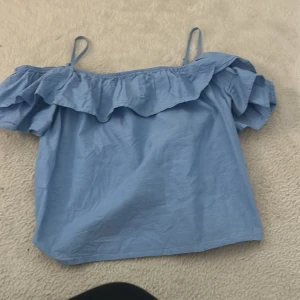 Ljusblå offshoulder topp med volang - Superfin ljusblå offshoulder topp med bred volang längs halsen och tunna axelband. Luftig och perfekt för varma dagar, med en söt och ungdomlig vibe. Materialet känns lätt och bekvämt, och volangen ger extra stilpoäng.