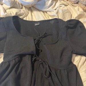 Svart kort klänning från Gina Tricot - Supersöt svart klänning från Gina Tricot med puffärmar och snygg snörning framtill. Den har en ribbad struktur och är tillverkad i mjukt material som sitter skönt. Passar perfekt om du gillar en feminin och trendig look med lite volym på axlarna.