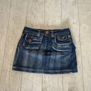 Lågmidjad mini jeanskjol - Snygg blå jeanskjol från bj33 med coola detaljer som stora fickor fram och bak, kontrastsömmar i rött och gult samt dekorativa knappar. Kjolen är kort och har en klassisk denimlook med slitningar och broderade detaljer baktill.Storlek 27 Midjemått 36 Längd 33