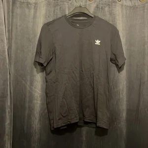Svart Adidas t-shirt med logga - Svart t-shirt från Adidas med klassisk vit treklöver-logga på bröstet. Enkel och stilren design med rund hals och korta ärmar. Perfekt för dig som gillar sportig och clean streetstyle.