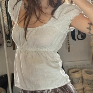  blus med puffärm - Supersöt  blus med tunna ränder och puffiga korta ärmar. Modellen har smock i midjan för en markerad siluett och knappar framtill. Perfekt för dig som gillar romantisk och vintageinspirerad stil. Tunt, mjukt bomullstyg som känns luftigt och skönt.