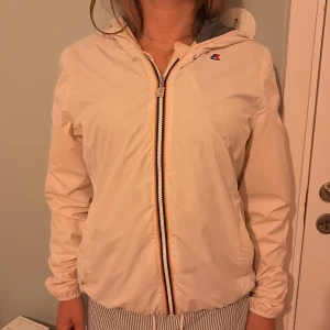  vindjacka från K-Way - Snygg vindjacka från K-Way med huva och klassisk randig dragkedja framtill. Jackan har en enkel, clean look och är perfekt för blåsiga dagar. Lätt material och elastiska muddar vid ärmslut. Liten K-Way-logga på bröstet.