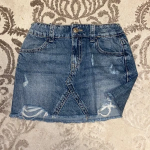 Blå jeanskjol från Gina Tricot - Snygg blå jeanskjol från Gina Tricot med slitna detaljer och fransad nederkant. Kjolen har klassiska fickor fram och bak, knapp och dragkedja i midjan samt coola sömmar som ger extra edge. Perfekt för dig som gillar en avslappnad och trendig stil. Andvänd 1 gång