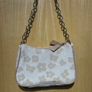 Beige blommig axelväska med kedja - Supersöt axelväska i beige och vitt med blommigt mönster. Väskan har en silvrig kedjerem och dragkedja med läderdetalj. Perfekt storlek för mobilen och det viktigaste. 