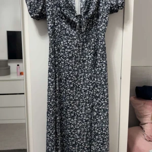 Blommig långklänning från SHEIN - Supersöt långklänning från SHEIN med mörkblå botten och vita blommor. Klänningen har korta puffärmar, v-ringning med knyt och dragkedja i ryggen. Materialet är mjukt och lätt, perfekt för varma dagar. Passformen är normal och klänningen har en fin slits framtill.