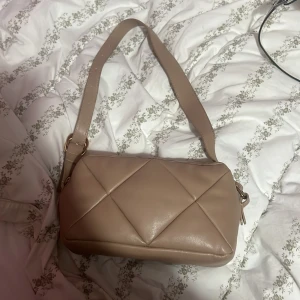 Beige quiltad axelväska i skinnimitation - Säljer en beige axelväska med quiltad framsida och bred axelrem. Väskan har en stilren, rektangulär form och stängs med dragkedja. Perfekt för dig som gillar minimalistisk och trendig design.
