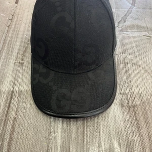 Svart keps från Gucci i ull och kashmir - Snygg svart keps från Gucci med diskret GG-mönster över hela kepsen. Tillverkad i en lyxig blandning av merinoull och kashmir. Klassisk form med böjd skärm och justerbar rem bak. Perfekt för dig som vill ha en stilren och exklusiv accessoar.