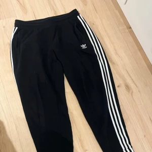 Svarta Adidas mjukisbyxor med ränder - Svarta mjukisbyxor från Adidas med klassiska vita tre ränder längs sidorna och broderad logga på låret. Byxorna har resår i midjan och är gjorda i mjukt bomullsmaterial. Perfekta för chill eller träning.