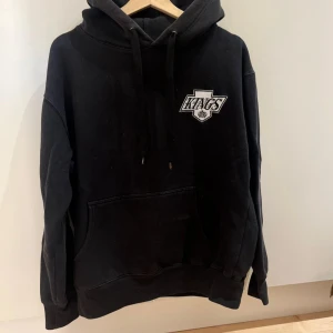 Svart hoodie Nucca Kings L - Svart hoodie från Nucca by Carl Thomas King i storlek L. Stor Kings-logga på ryggen och mindre logga på bröstet. Klassisk känguruficka, snörning i huvan och mjukt material. Perfekt för dig som gillar streetwear och sportig stil.