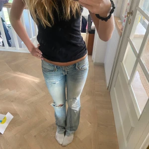 Bootcut jeans från American Eagle - Snygga ljusblå bootcut jeans från American Eagle med slitningar och hål på knäet. Jeansen har låg midja, klassisk femficksdesign och dragkedja. Perfekt för dig som gillar en avslappnad och trendig look. Jeansen är sydda till bootcut och har även en tillsydd fel längst ner, men tycker inte att det är något man tänker på när dom sitter på 