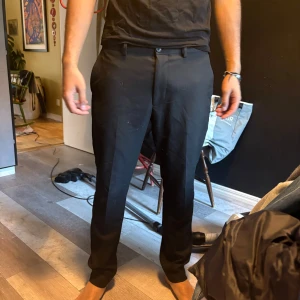 Svarta kostymbyxor Riley Slim Fit - Svarta kostymbyxor från Riley i slim fit-modell. Byxorna har klassisk rak passform, pressveck och bälteshällor. Snygga och stilrena, perfekta till skjorta eller t-shirt. Materialet är slätt och känns dressat.