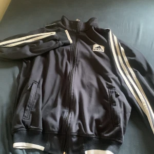 Svart Adidas vindjacka med vita ränder - Svart Adidas vindjacka med klassiska vita ränder längs ärmarna och dragkedja framtill. Jackan har hög krage, två fickor med dragkedja och ribbade muddar med vit detalj. Perfekt för en sportig och avslappnad stil. Storlek M/S. Pris kan diskuteras vid snabb affär